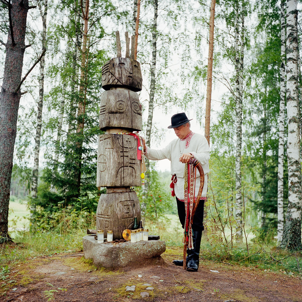 Aarne Leima, Ülembsootska (Viceroy in Seto language) from 2013-2014 posing with a representation of Peko, his king on the hill of the gods of Kolossova. Historically Peko is also the fertility god. In the past, villages of Setomaa had a statuette stored in villager weakest man's barn. Peko then brought him strength, help and luck. Each fall after harvest, the village men fought themselves. The first one to bleed, earned the right to be protected by Peko. Now, since the restoration of Estonian independence and division of Setomaa by the Russian-Estonian border the Seto made Setomaa their kingdom and Peko their king. Since then, it is said that Peko is resting in the Petseri monastery caves in Russia. He will wake up only to defend the Seto from a great danger. His representative, the Ülembsootska, communicate with Peko through his dreams and must pass messages to the Seto people. The Ülembsootska is elected annually by the Seto during the day of the Kingdom, Kuningriik päev. There are various legends about the hill gods Kolossova: there, God would take souls to lead them to heavens by putting them on a carriage. There would be a departure once a year. From this hill it was possible to see the domes of the Petseri Monastery. Believers used to come and pray, bowing to the monastery. Kolossova, Setomaa, Estonia. 1st August 2014.