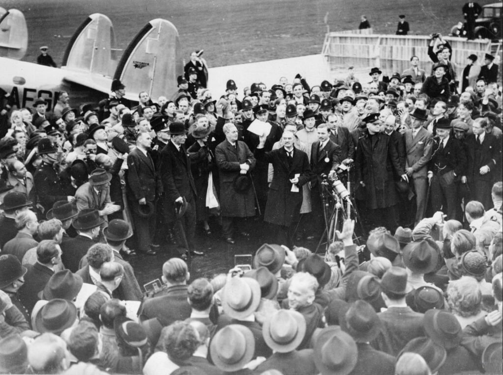 Neville Chamberlain showing the Anglo-German Declaration to commit to peaceful methods signed by both Hitler and himself, at Heston Aerodrome, on his return from Munich on 30 September 1938. Public domain.
